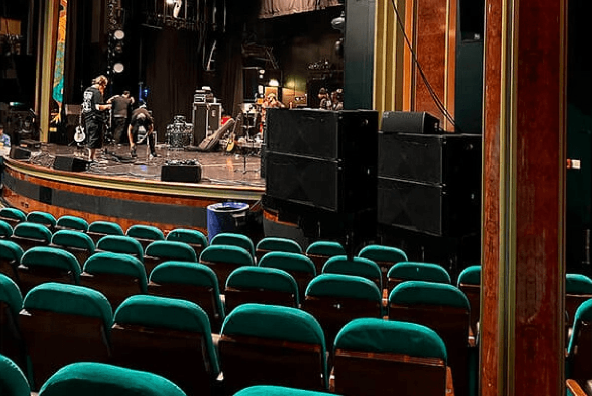 Stage crew setting up guitars and equipment on a theater stage, with rows of empty green seats in the foreground (auditorium).