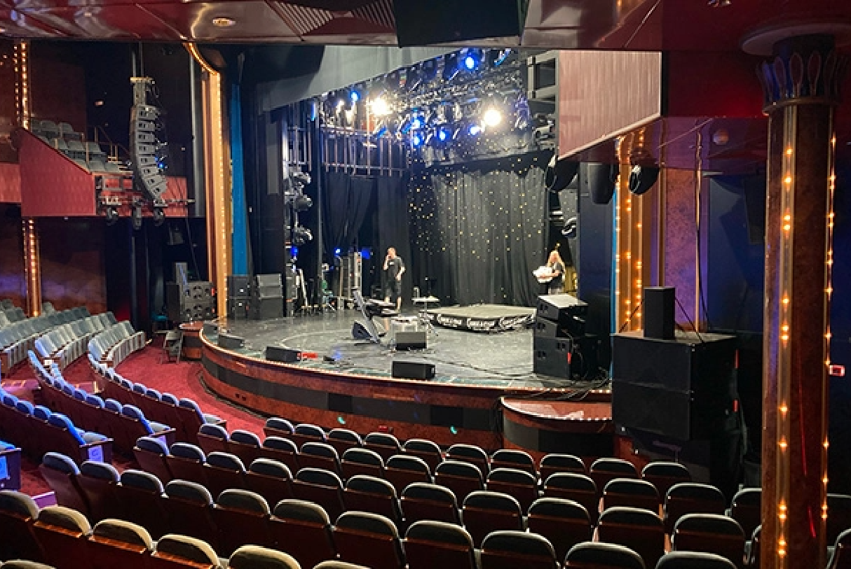 Theater stage with performers and equipment under blue/white lights; empty red seats extend toward a dark backdrop with curtains and stage gear nearby.