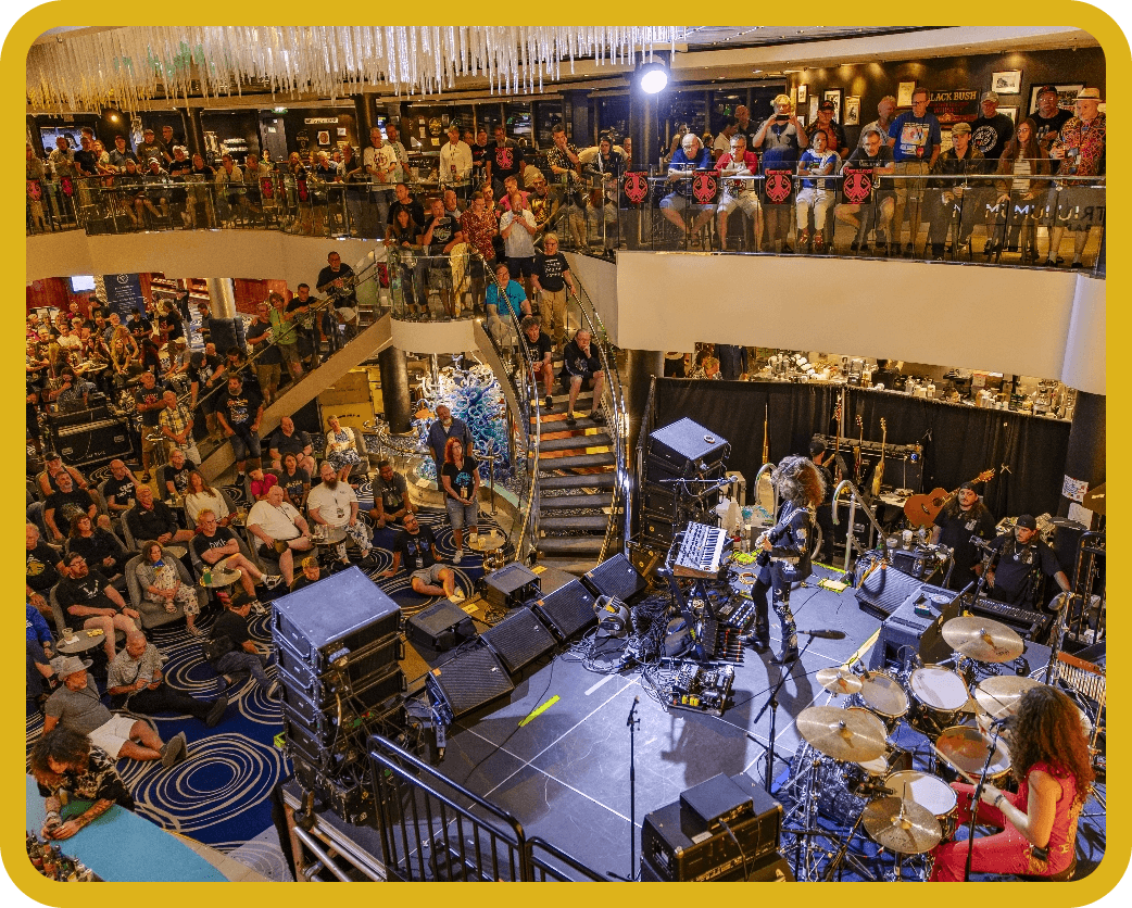 Live band performing on a stage inside a multi-level atrium with a crowd on ground and balcony rooft, drums and guitarists visible.