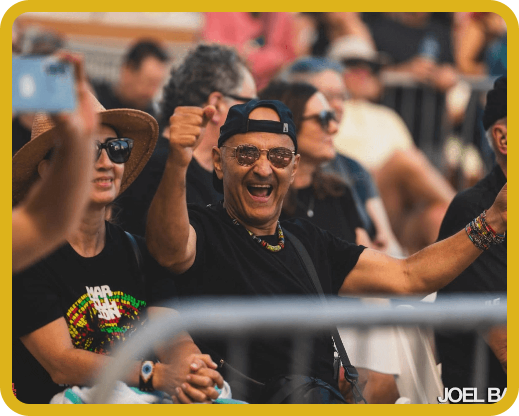 Crowd at an outdoor event cheering, with a man in a cap and sunglasses raising his fist.