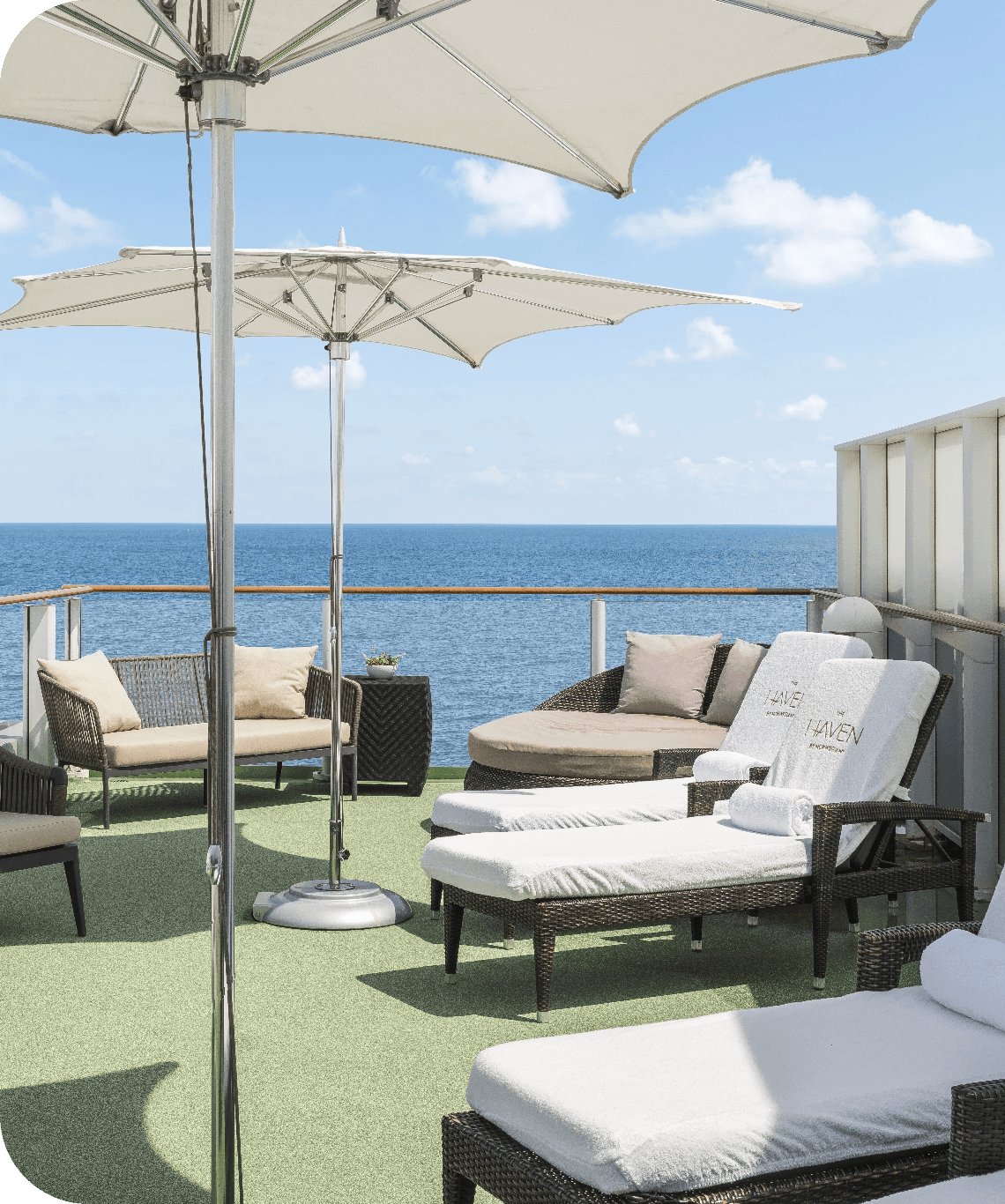 Rooftop terrace with wicker lounge chairs, beige cushions, and large white umbrellas overlooking the ocean.