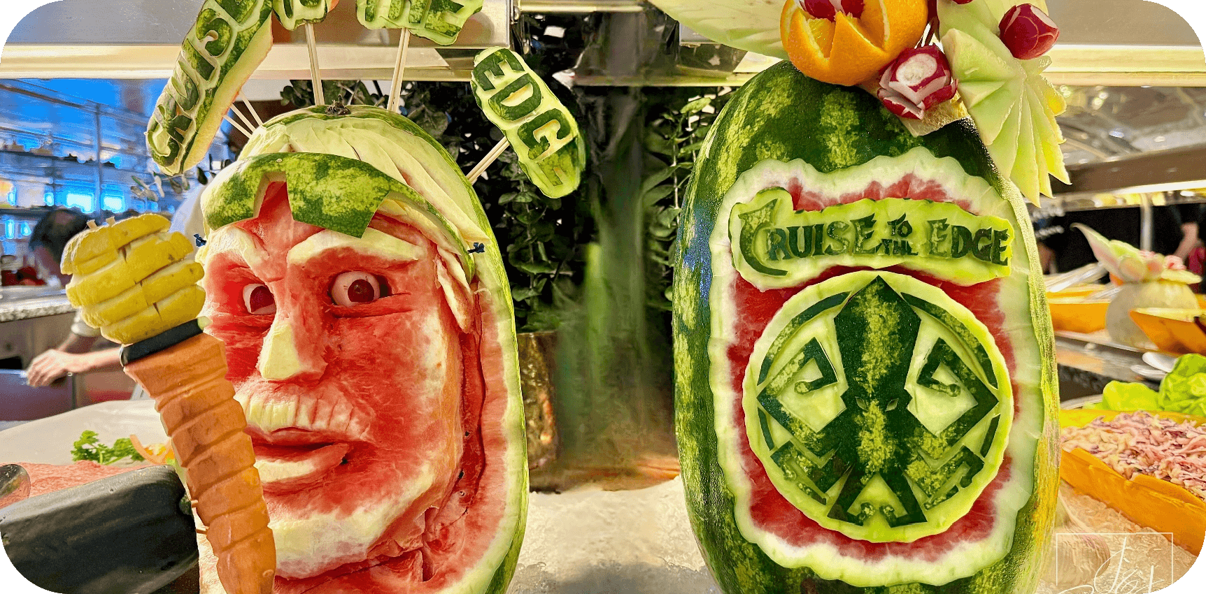 Two carved watermelons with cartoon faces and fruit garnishes on a buffet display, resembling a fun fruit sculpture showpiece.
