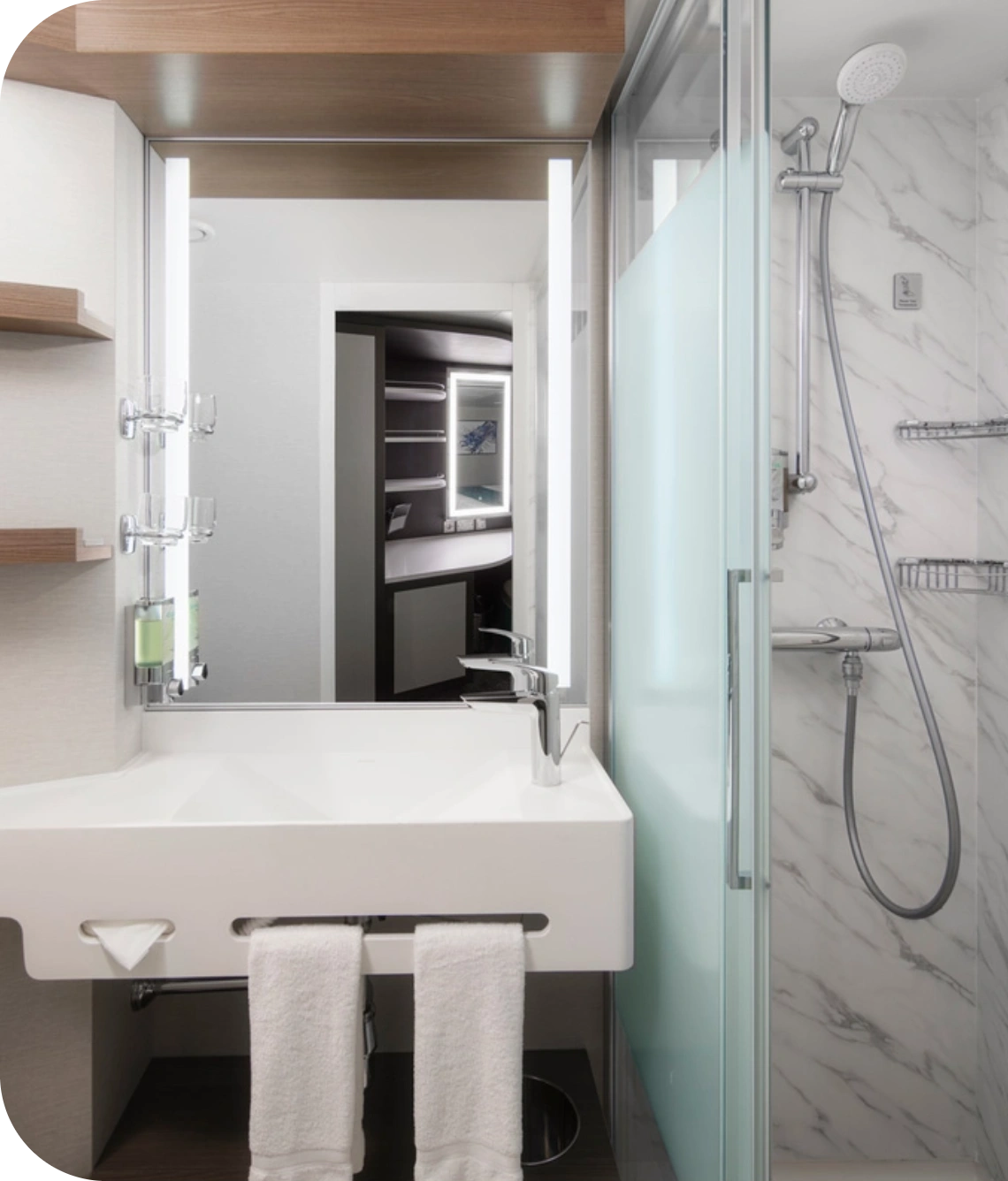 Modern bathroom with a white rectangular sink, chrome faucet, illuminated mirror, and a glass shower enclosure to the right; two white towels hang beneath the sink.
