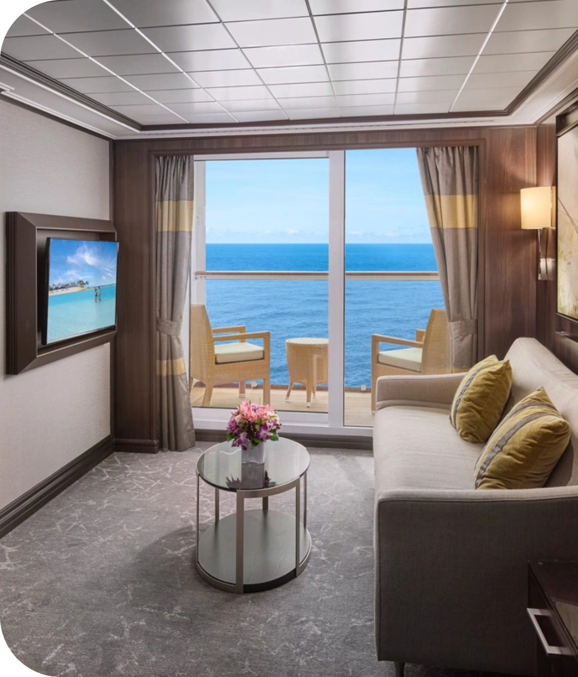 Hotel living room with ocean view through sliding glass doors; sofa, glass coffee table with flowers, and wall-mounted TV on the left; balcony seating beyond.