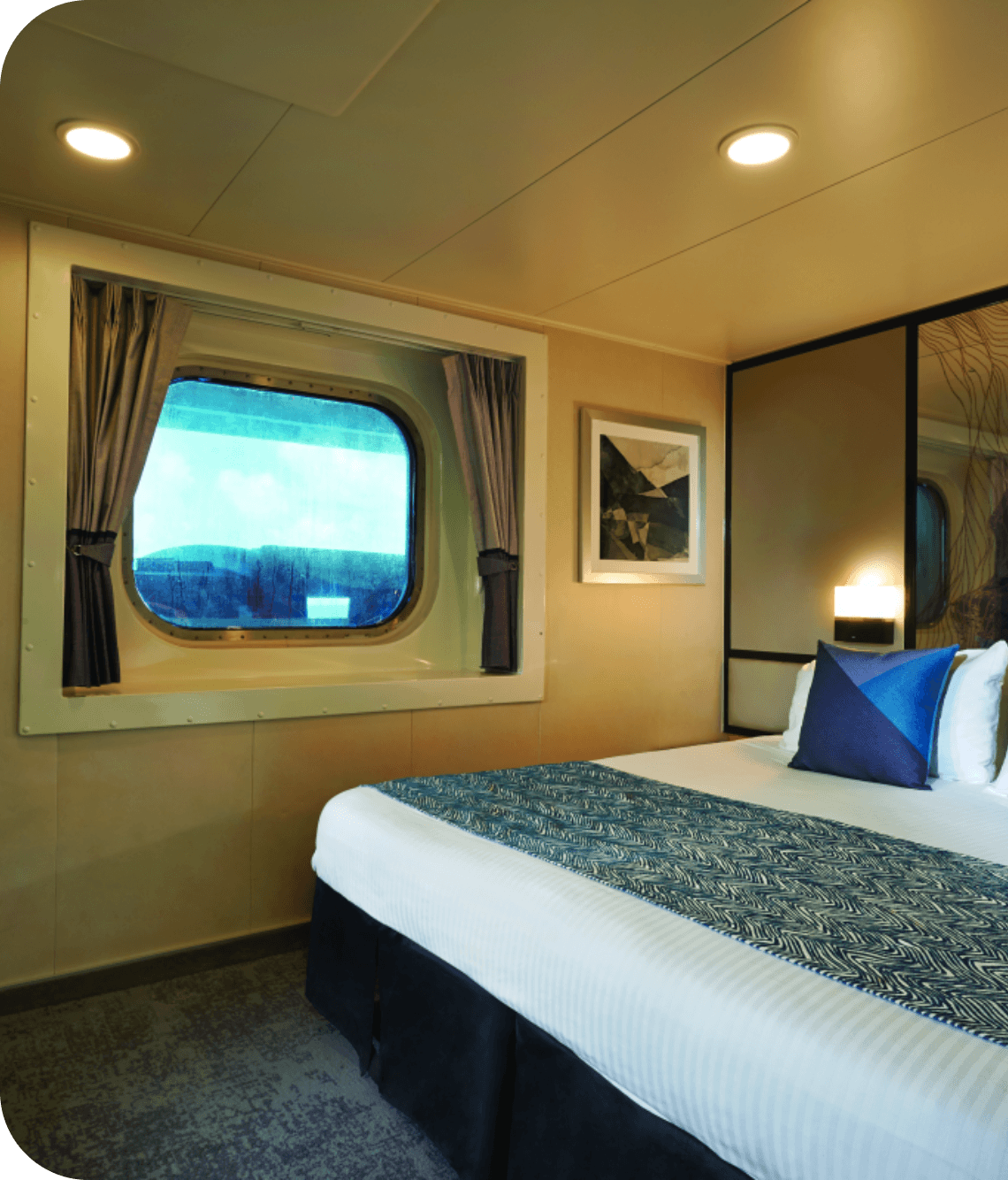 Hotel/ship cabin bedroom with a round porthole window, curtains, framed geometric art, and a neatly made bed with blue pillows and a decorative runner.
